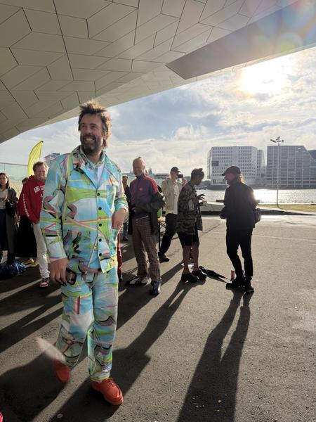 A lively outdoor scene unfolds at the IJpromenade in Overhoeks, Amsterdam, where a man in a vibrant, patterned suit smiles brightly while walking